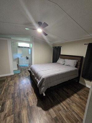 Een slaapkamer met een bed, een plafondventilator, een raam en een badkamer in de achtergrond.
