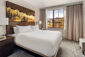 Habitación de hotel con una cama grande, una mesita de noche, una lámpara, un cuadro en la pared, una ventana con cortinas y vistas a edificios.