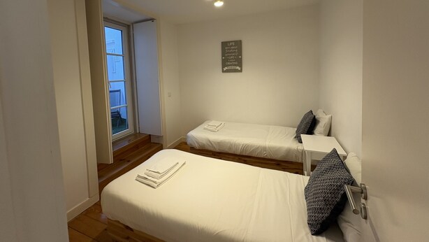 A small, clean room with two beds, a small table, a wall-mounted light fixture, and a door leading to a balcony. - Alfama Premium (Lisbon)