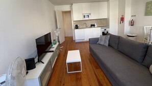 A modern living space with a flat-screen TV, a white coffee table, a grey sofa, a kitchen area with white cabinets, a microwave, and a small dining table. - Alfama Premium (Lisbon)