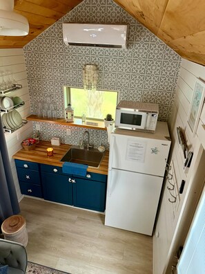 A compact kitchen with a microwave, sink, and a refrigerator. There is a window above the sink, and a patterned backsplash.