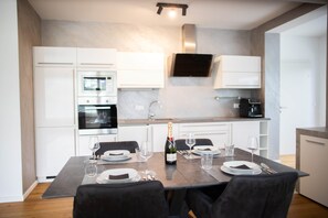 A modern kitchen with white cabinets, a built-in oven, and a dining table set for two with wine glasses and a bottle of champagne.