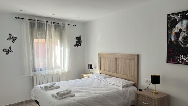 Et soveværelse med seng, et natbord, en lampe, en radiator og et vindue med let gardin.