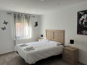 A bedroom with a bed, a nightstand, a lamp, a radiator, and a window with sheer curtains.