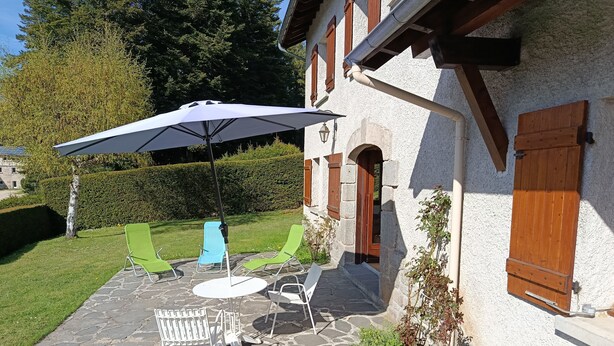 Een terras van natuursteen met tuinmeubilair, een grote parasol en een gebouw met houten luiken.