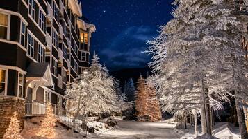 A snow-covered pathway leading to a building with multiple windows and balconies, surrounded by snow-laden trees.