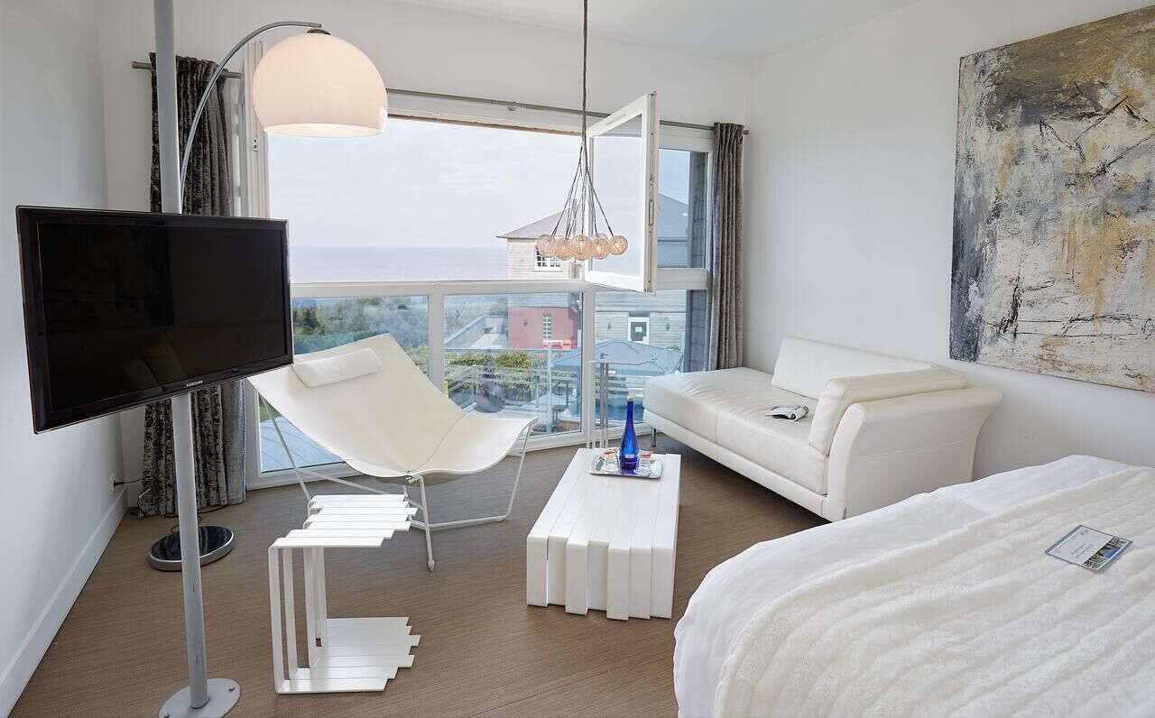 A modern hotel room with a large window, a flat-screen TV, a white sofa, a white armchair, a white coffee table, and a bed.
