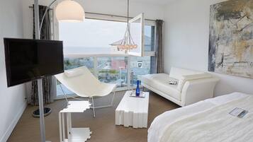 A modern hotel room with a large window, a flat-screen TV, a white sofa, a white armchair, a white coffee table, and a bed.