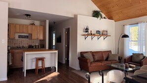 Una cocina moderna con armarios de madera, un taburete de bar y una mesa de comedor. Hay una sala de estar con un sofá de cuero marrón, una mesa de centro de vidrio y una lámpara.
