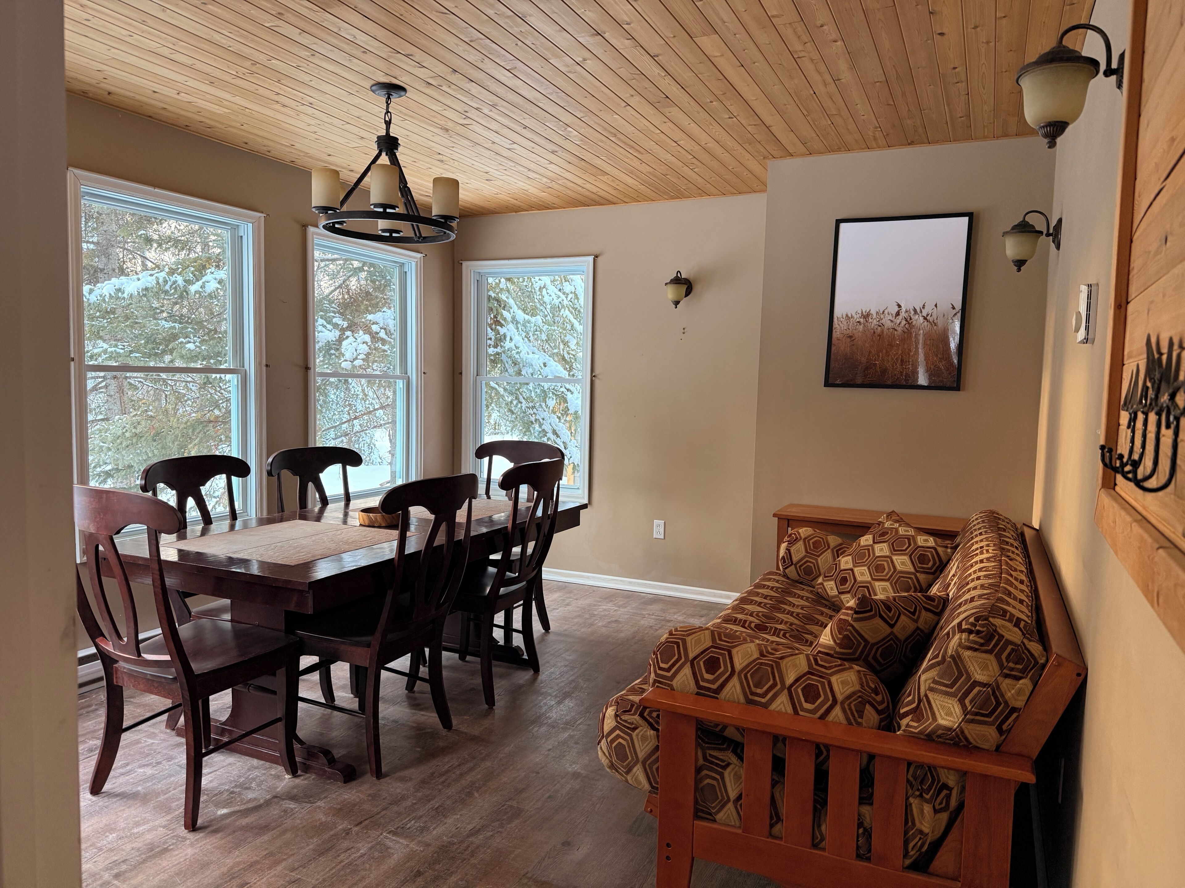 Un comedor con mesa y sillas de madera, techo de madera y ventanas amplias con vista a árboles nevados.
