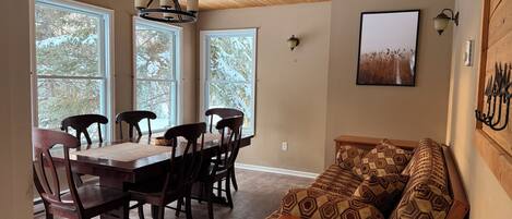 Un comedor con mesa y sillas de madera, techo de madera y ventanas amplias con vista a árboles nevados.