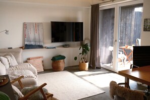 Ein Wohnzimmer mit einer Couch, einem Fernseher, einem Esstisch und einer Glastür, die zu einem Außenbereich führt.