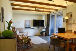 Ein Wohnzimmer mit einer Couch, Sesseln, einem Esstisch, einem Fernseher und einem Fenster mit Vorhängen.