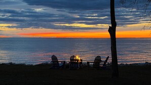 Een strandlandschap met drie Adirondack stoelen en een vuurkuil, met uitzicht op een zonsondergang boven de oceaan.