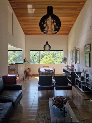 A spacious living area with a wooden ceiling, large windows with a view of greenery, a dining table with chairs, a sofa, and a coffee table with flowers.