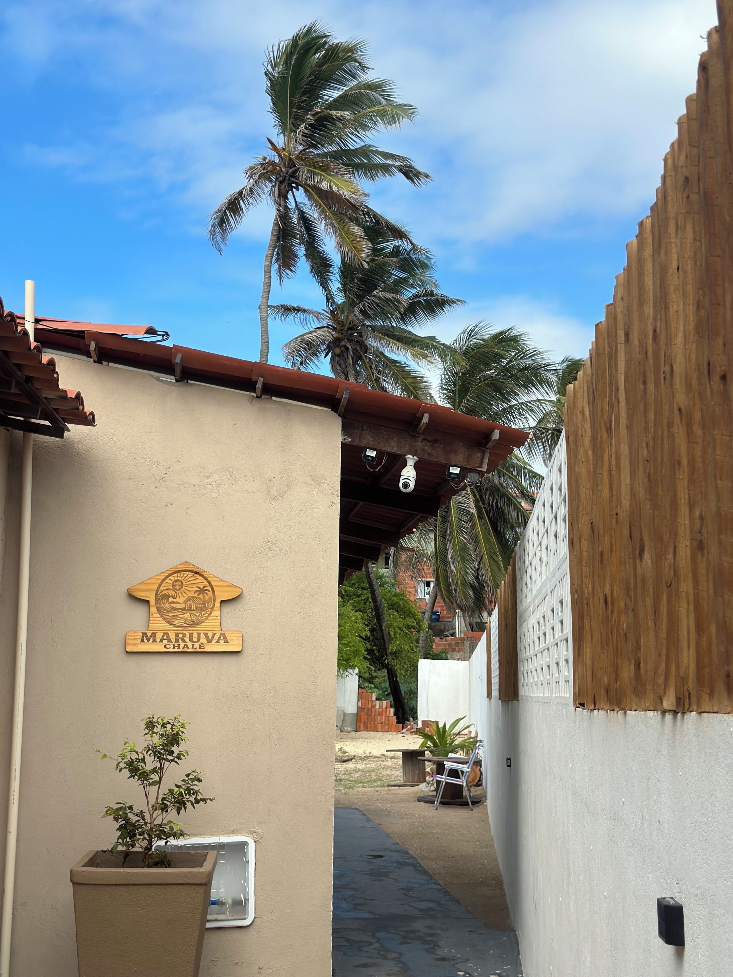 Um beco estreito com cerca de madeira, uma palmeira e um edifício com uma placa que diz 'MARUVA CHALE'.