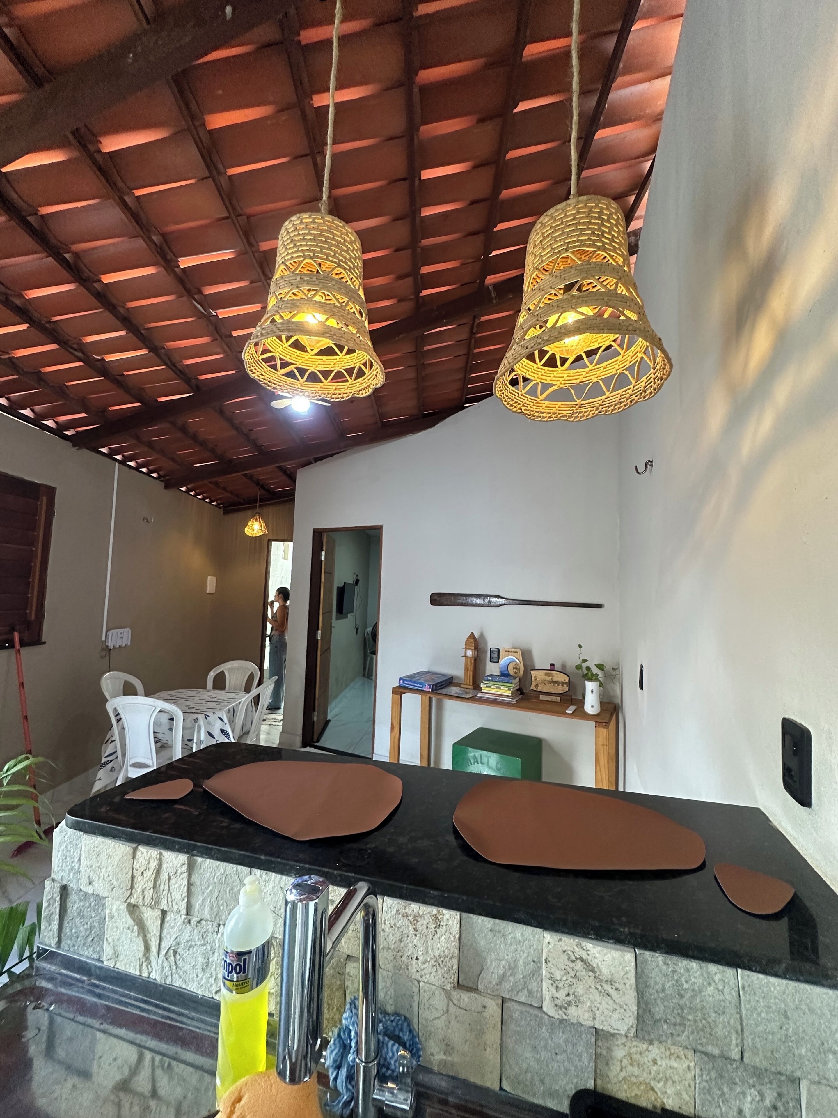Uma cozinha com bancada de pedra, duas luminárias de vime suspensas, uma bengala de madeira na parede e uma pequena mesa com livros e uma planta.