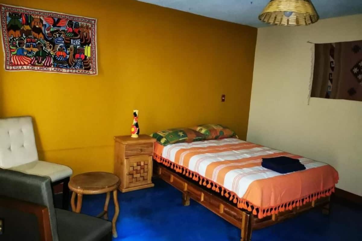 A room with a bed, a chair, a small table, a wall hanging, and a hanging light fixture.