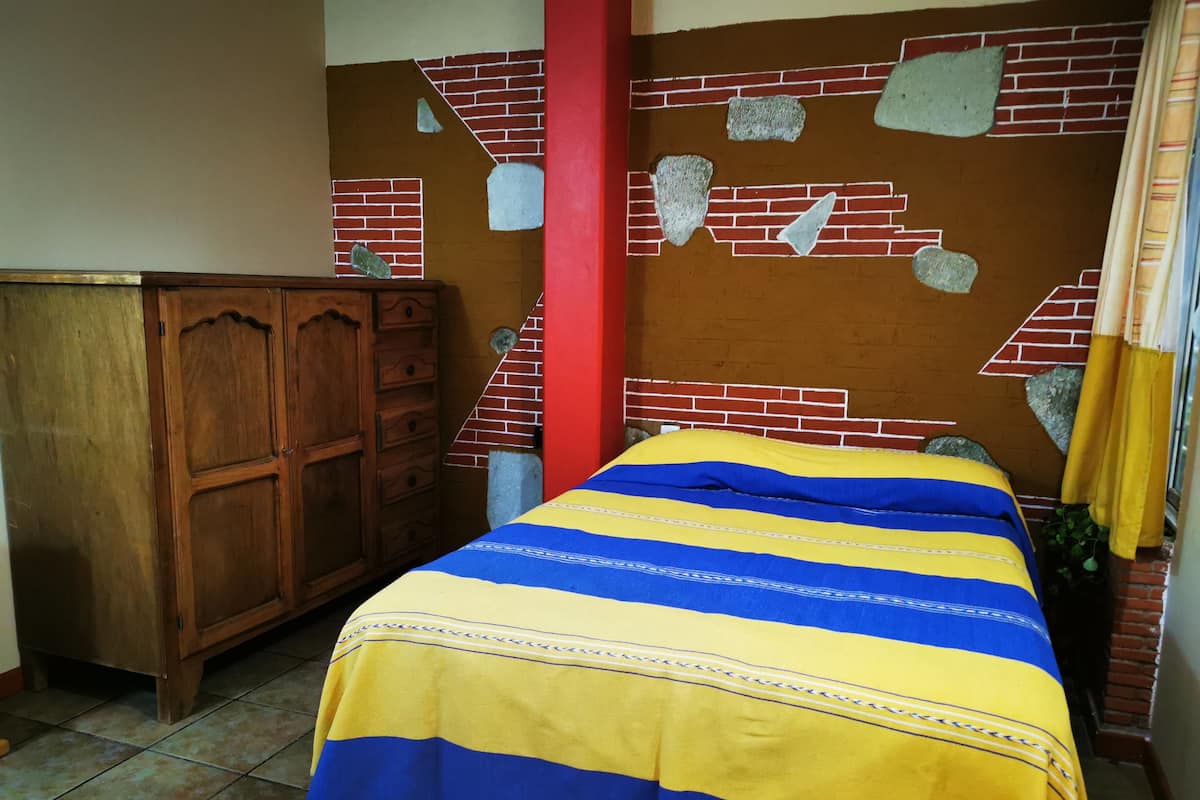 A room with a bed, a wooden dresser, and a brick wall with a red pillar.