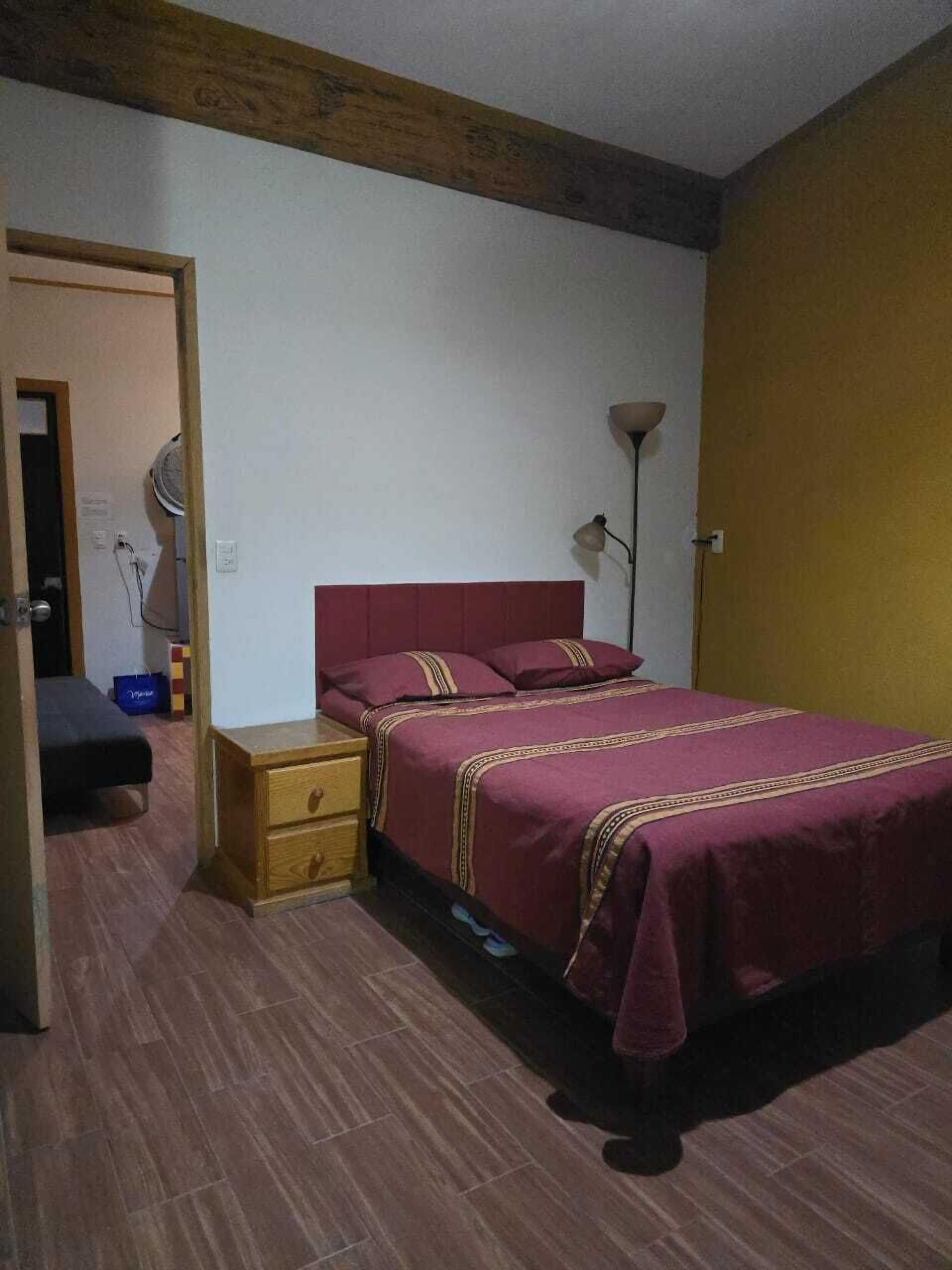 A bedroom with a wooden headboard, a single bed with a red bedspread, a bedside table with drawers, a floor lamp, and a door leading to another room.