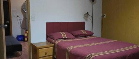 A bedroom with a wooden headboard, a single bed with a red bedspread, a bedside table with drawers, a floor lamp, and a door leading to another room.