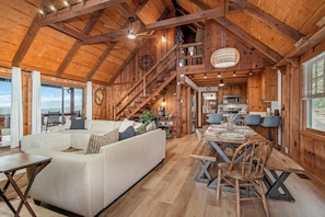 A spacious living area with a large sofa, wooden dining table, and a staircase leading to an upper level. - Views • Hot Tub • Fenced Yard • Near Downtown (Blue Ridge)