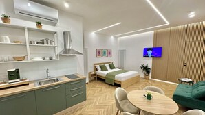 Habitación de hotel moderna con kitchenette, cama con edredón verde, televisor montado en la pared y zona de comedor con mesa y sillas redondas.