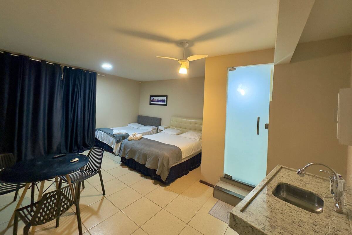 Habitación de hotel con dos camas, una mesa de comedor, sillas, un ventilador de techo, un baño con lavamanos y un cuadro en la pared.