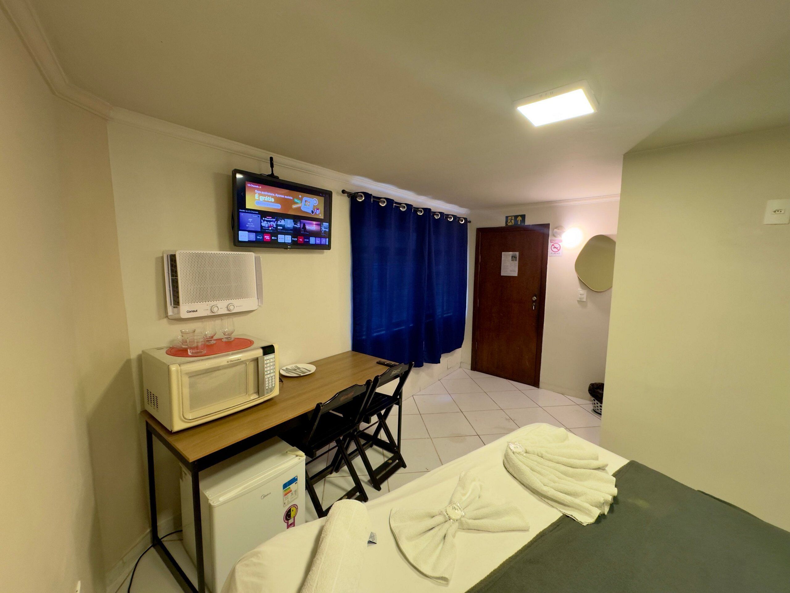 A small room with a bed, a desk with a computer, a TV, a microwave, and a small refrigerator.