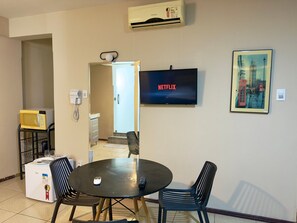 A room with a dining table, chairs, a microwave, a refrigerator, a TV displaying 'NETFLIX,' and a painting of a cityscape.