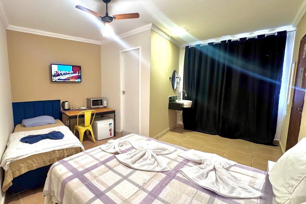 Habitación de hotel con dos camas, televisor, escritorio con computadora y microondas, refrigerador pequeño y baño con lavamanos y espejo.