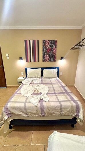 A neatly made bed with a checkered bedspread, two white pillows, and a white bathrobe on it. There are two wall-mounted light fixtures and two paintings above the bed.