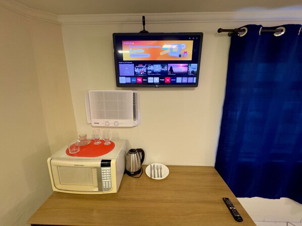 A hotel room with a television mounted on the wall displaying a channel guide, a white bedside table with a microwave, a kettle, and a remote control.