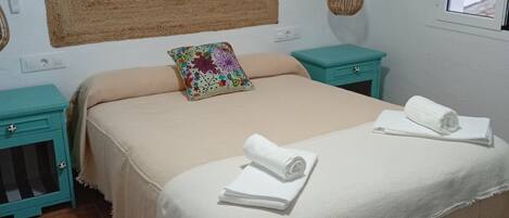 Un lit recouvert d’une couverture beige, une serviette enroulée et un coussin coloré. On y trouve deux tables de chevet turquoise avec des tiroirs, une tête de lit en osier et un panier suspendu.