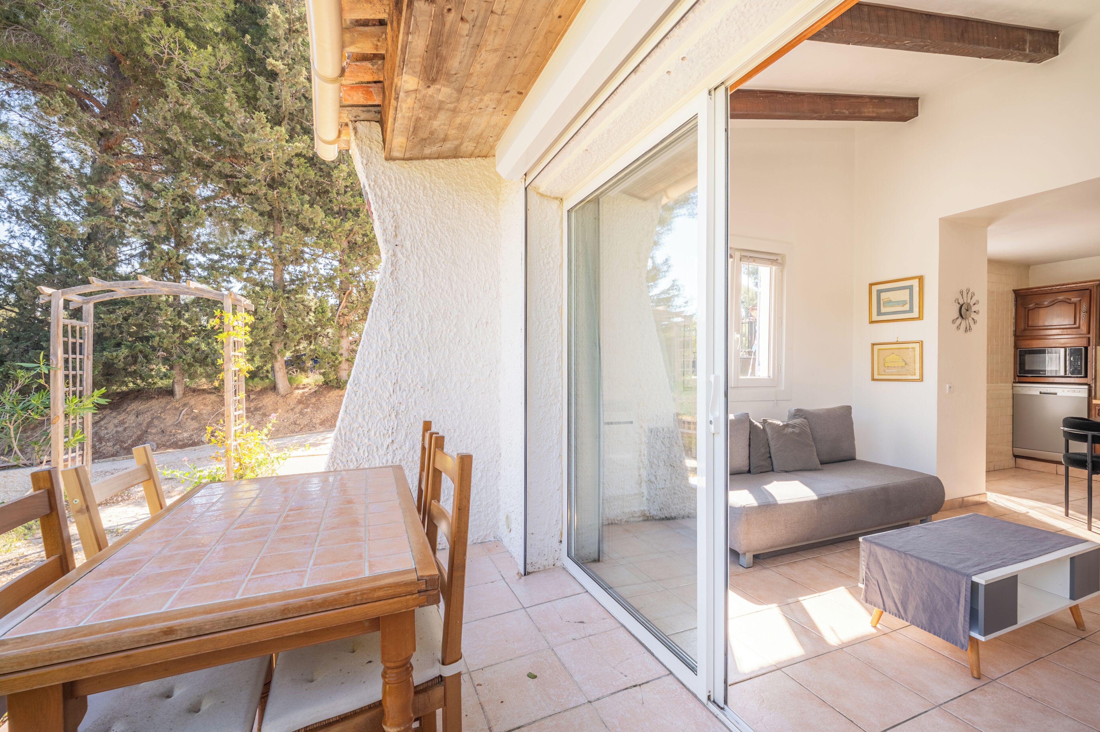 Een kamer met een houten tafel en stoelen, een bank, een klein poufje en een schuifdeur van glas die leidt naar een buitenruimte met een pergola en bomen.