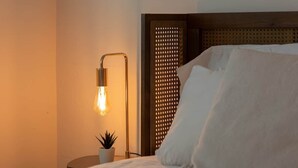 A bed with white linens, a bedside lamp, a small potted plant, and a decorative wall panel. - Mid-Century Oasis w/ heated Pool (Tucson)