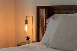 A bed with white linens, a bedside lamp, a small potted plant, and a decorative wall panel. - Mid-Century Oasis w/ heated Pool (Tucson)