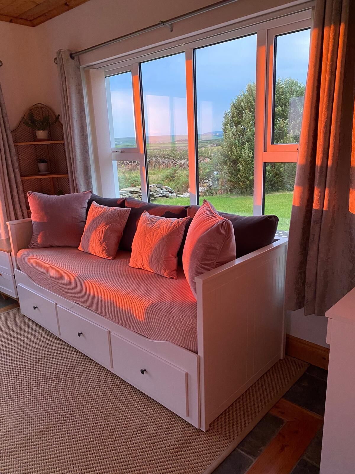Ein weißes Sofa mit Stauraum, große Fenster mit Vorhängen und Blick auf eine grüne Landschaft.