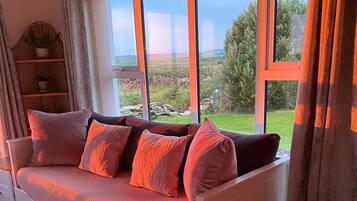 Ein weißes Sofa mit Stauraum, große Fenster mit Vorhängen und Blick auf eine grüne Landschaft.