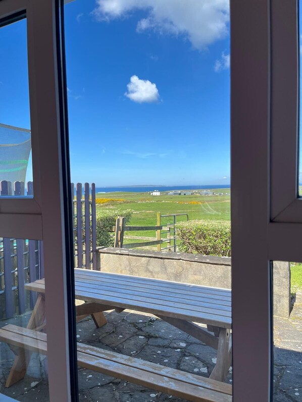 Blick durch ein Fenster auf eine mit Stein gepflasterte Terrasse, Holzbanken und einen Zaun mit Blick auf ein saftiges Grasfeld und entfernte Gebäude.