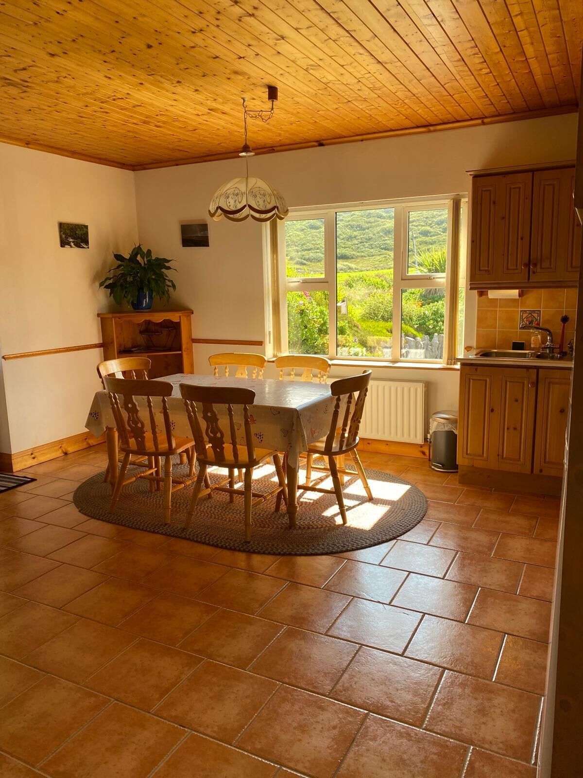A dining area with a round table and chairs, a window with a view of greenery, wooden ceiling, and a kitchen area with cabinets.