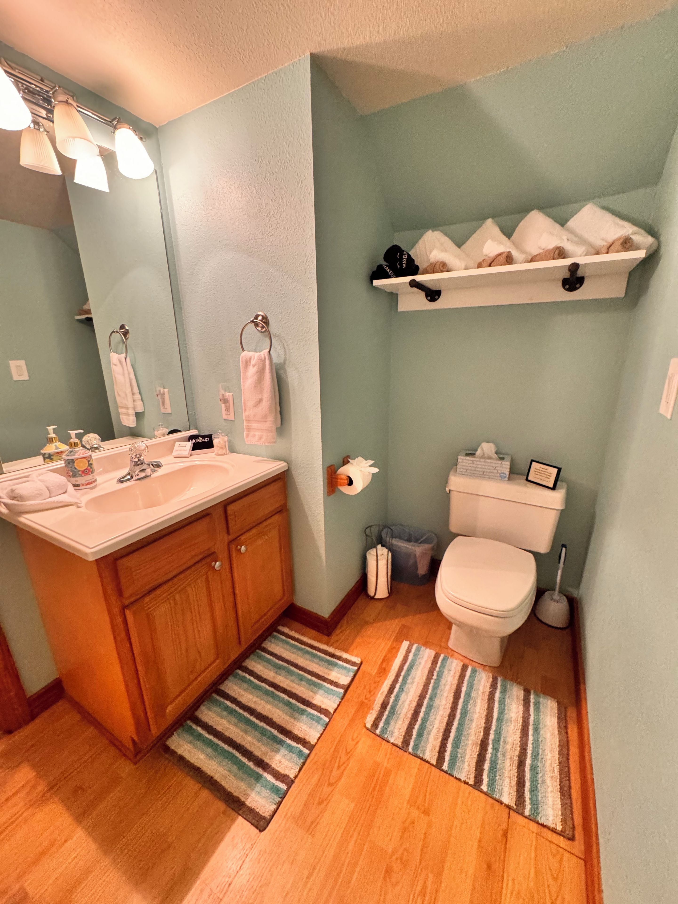 A bathroom with a wooden vanity, a toilet, a shelf with towels, and a striped rug.