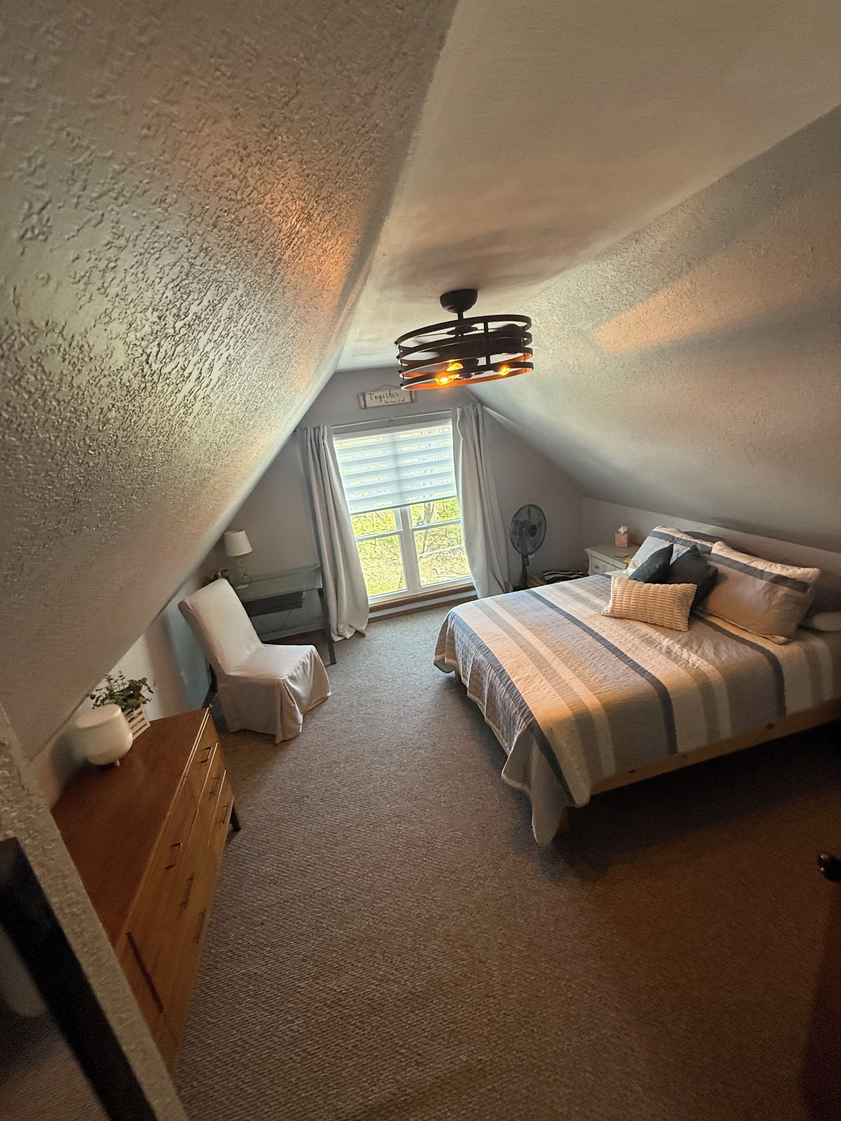A bedroom with a sloped ceiling, a bed with a striped bedspread, a bedside table, a chair, a window with curtains, and a ceiling light fixture.
