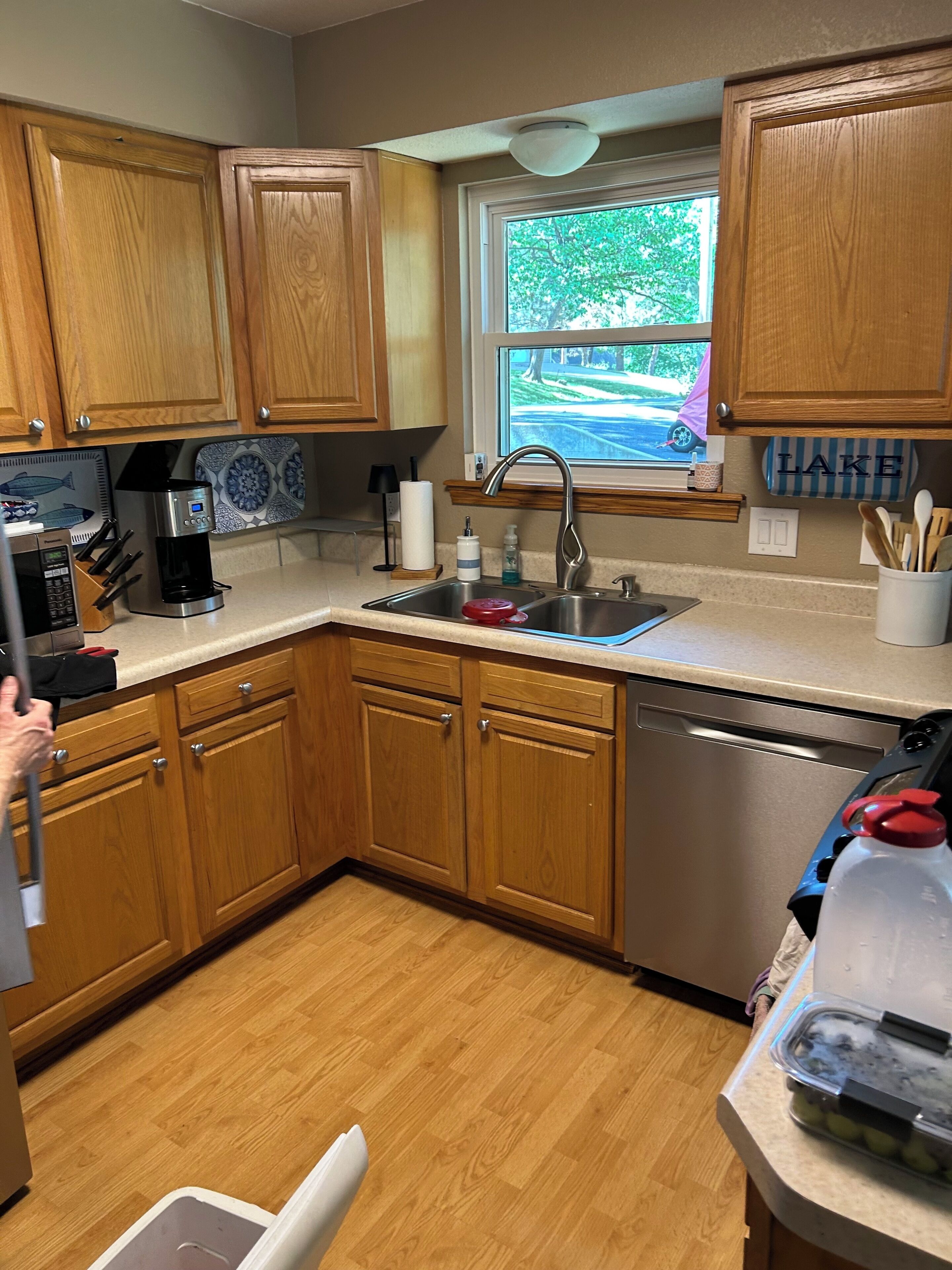 A kitchen with wooden cabinets, a stainless steel sink, a coffee maker, and a window with a view of a road and trees.