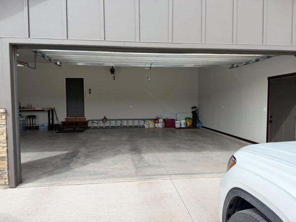 A garage with a white car parked inside, a workbench with tools, and various items on the floor. - Rally Rider's Retreat! Paved Roads, large driveway & Centrally located (Spearfish)
