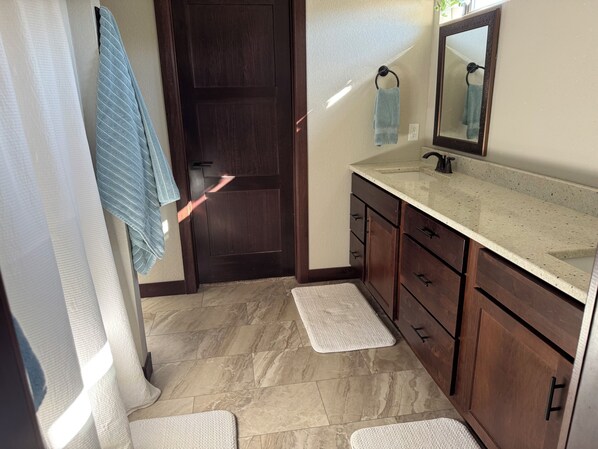 A bathroom with a dark wooden door, a white sink with a mirror, and two white mats on the floor. - Rally Rider's Retreat! Paved Roads, large driveway & Centrally located (Spearfish)