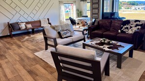Een moderne woonkamer met een bank, fauteuil en salontafel, een eethoek met tafel en stoelen, en een groot raam met uitzicht op het buiten.