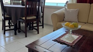 A dining area with a table and chairs, a sofa with a cushion, and a coffee table with a fruit bowl. - Θάλασσα Thea (Kavala Paleo tsifliki)
