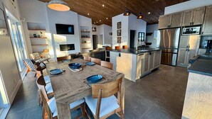A modern kitchen with a central island, built-in appliances, and a dining area with a long table and chairs. - Chalet 2025 sur lac Barron navigable Spa, quai, 4 rooms -14 pers - Laurentians (Gore)