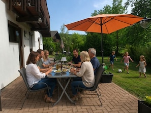 Eine Gruppe von Menschen speist an einem Tisch im Freien, beschattet von einem Sonnenschirm, umgeben von Pflanzen.